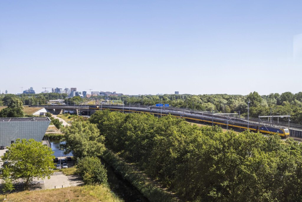 Uitzicht op de Thomas R. Malthusstraat in Amsterdam met een passerende intercitytrein en omliggend groen.